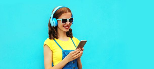 Portrait of happy smiling young woman in headphones listening to music with phone on blue background - Photo, image