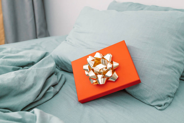 Red Christmas gift box present with gold bow on a grey bed in morning in bedroom. Valentines Day, Womens Day. Shopping concept and black Friday composition. - Photo, Image