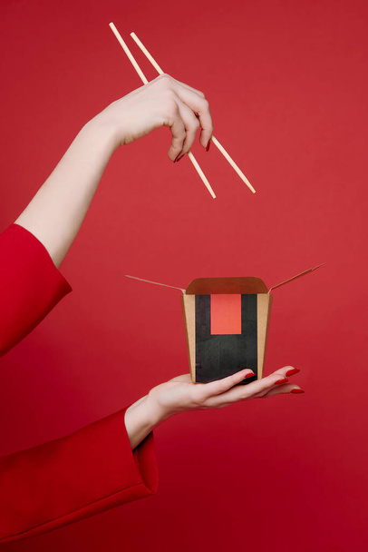 Woman's hands with chinese food in the box and wooden sticks on red background. Noodles and rise. Minimalism. Red clothes. Beige skin. Advertisement. Delivery.  East culture. Asian kitchen.  - Φωτογραφία, εικόνα