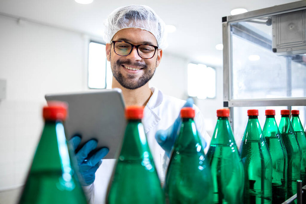 Doświadczony pracownik technologiczny w białym fartuchu laboratoryjnym i siatce do włosów kontrolującej produkcję butelkowanej wody oczyszczonej w fabryce butelkowania. - Zdjęcie, obraz