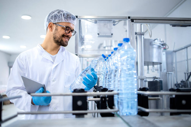 Technik przemysłowy trzymający tablet i sprawdzający produkcję wody butelkowanej w rozlewni na automatach. - Zdjęcie, obraz