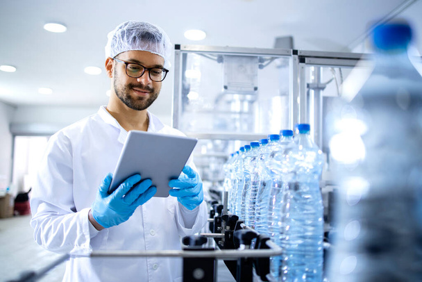 Pracownik zakładu butelkowania lub technik w sterylnej odzieży stojący przy automatycznej maszynie taśmowej i sprawdzający produkcję wody. - Zdjęcie, obraz
