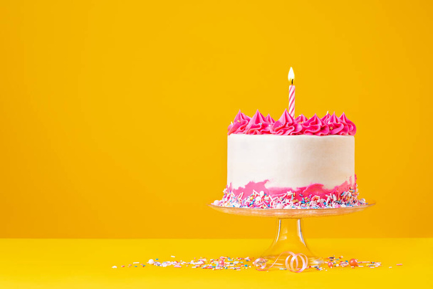 ピンクのバタークリームアイシング、カラフルなスプリンクル、明るい飽和黄色の背景にライト付きの誕生日キャンドル付きの誕生日ケーキ. - 写真・画像