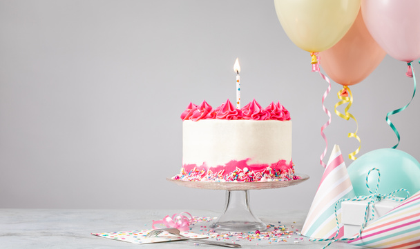 Pink Birthday cake with candle, presents, hats and colorful balloons over light grey background. Birthday Party! - Zdjęcie, obraz