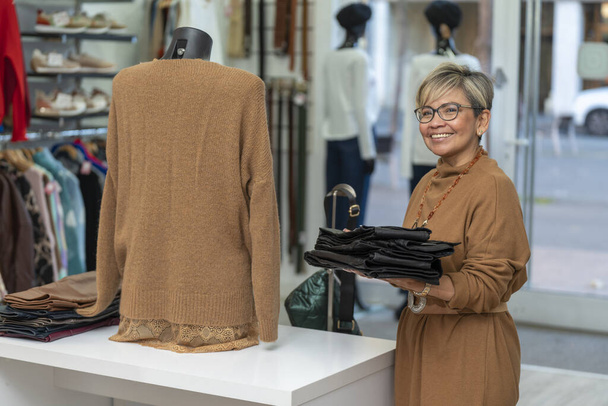 Senior latin vrouw kleding winkel bediende op zoek naar camera - Foto, afbeelding