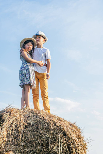 Retrato vertical de un niño y dos niñas que se quedan y abrazan firmemente, en un pajar en el campo. Niños con sombreros de paja. - Foto, imagen