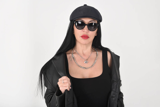 A stylish woman in the studio on a white background wearing sunglasses, jeans, a black T-shirt, a black cap and a black leather jacket in retro style - Fotoğraf, Görsel