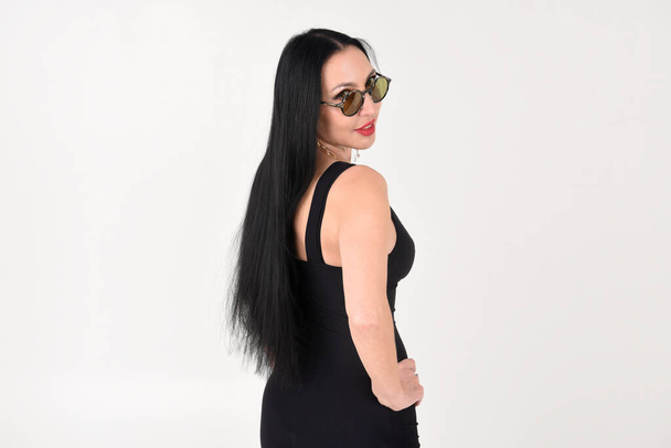 The brunette girl is beautiful and stylish in a black dress and dark glasses with her feet in the studio on a white isolated background - Zdjęcie, obraz