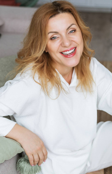 Krásná blondýnka středního věku s rozzářeným úsměvem sedící doma vedle pohovky a dívající se do kamery. Fotografie - Fotografie, Obrázek