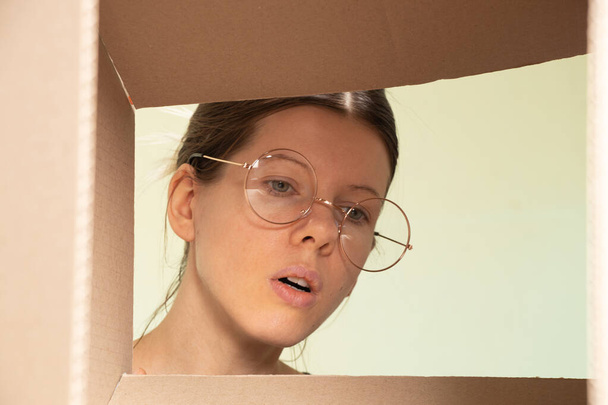 Το πρόσωπο του κοριτσιού κοιτάζει μέσα στο χαρτόκουτο, παραλαμβάνει το δέμα, κοιτάζει μέσα από το κουτί. - Φωτογραφία, εικόνα