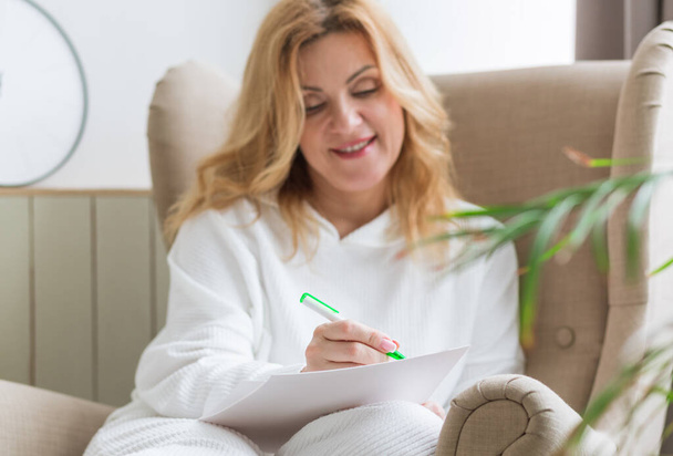 Una mujer vestida de blanco se sienta en una silla de casa y escribe sus sueños en papel. Concepto de visualización del deseo - Foto, Imagen