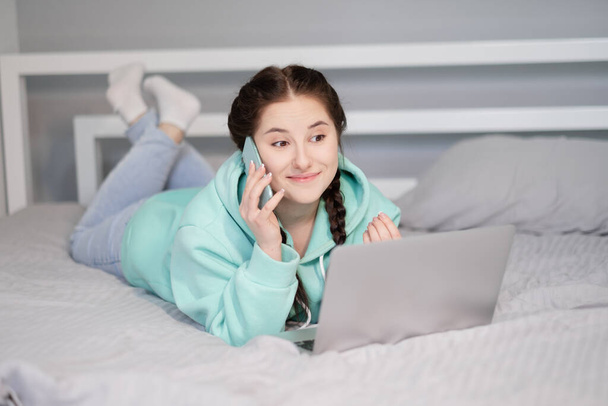 Distance learning online education and work. Happy brunette woman girl working office work remotely from home in turquoise hoodie and jeans on bed. Talking phone. Using computer laptop. - Φωτογραφία, εικόνα