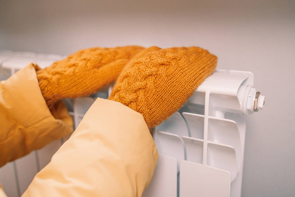 Frauenhände in gelben Strickhandschuhen berühren einen weißen Heizkörper im Haus. Temperaturkontrolle. - Foto, Bild