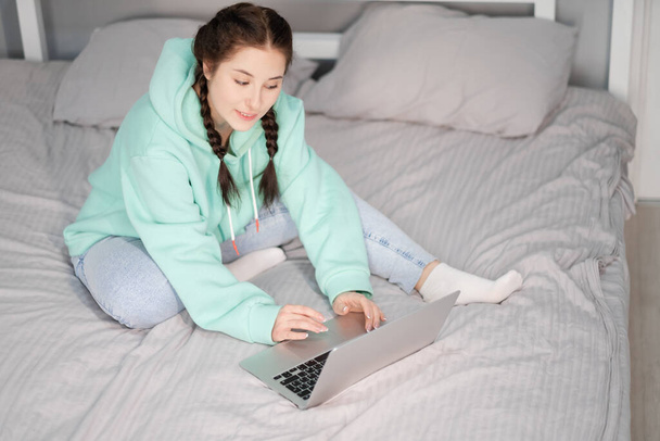 εξ αποστάσεως εργασία ή μακρινή μελέτη από το σπίτι. νεαρή μελαχρινή κοπέλα στο κρεβάτι με φορητό υπολογιστή. περιορισμοί καραντίνας. πανδημία του covid-19 - Φωτογραφία, εικόνα