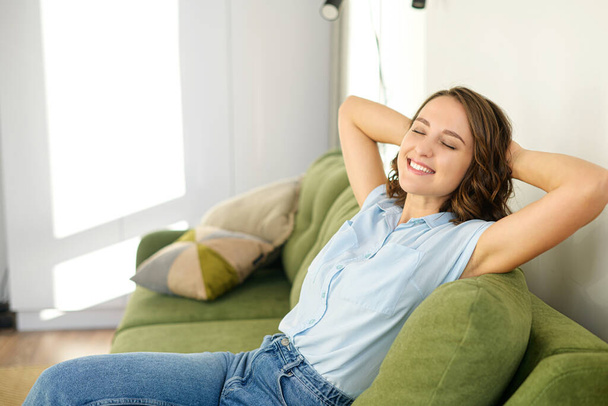 Portrait of charming woman with brunette hair. Smiling young female sitting on the couch and closed her eyes while relaxing - Zdjęcie, obraz
