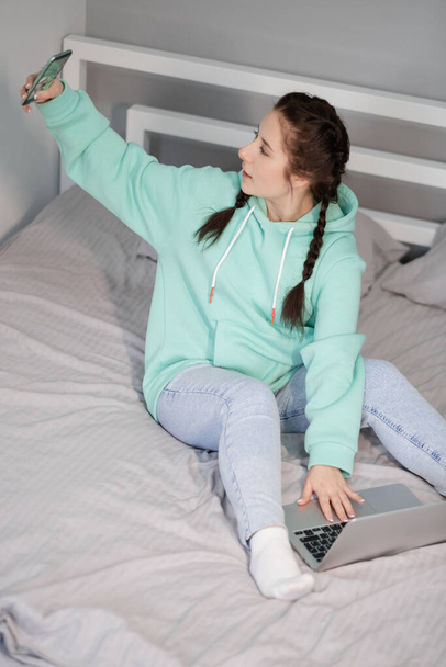 working or studying from home. young brunette woman in turquoise hoodie and jeans on bed with laptop and turquoise color phone. quarantine restrictions. distant work, study. happy remote education - Fotó, kép