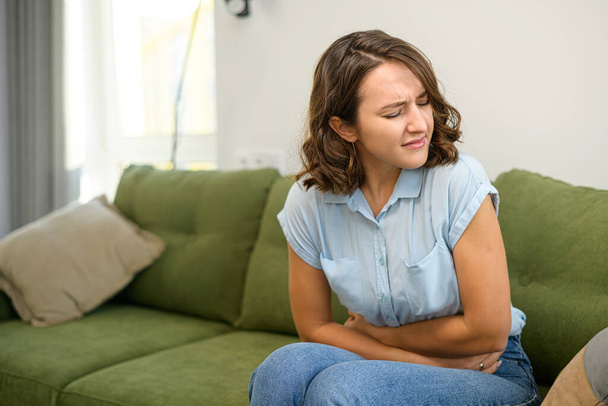 Young caucasian woman with curly hairstyle sitting at the sofa and suffering from stomach ache, keeping hands on her stomach at home, waiting for doctor - Foto, afbeelding