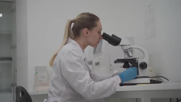 Γυναίκα επιστήμονας που χρησιμοποιεί μικροσκόπιο ενώ εργάζεται στο εργαστήριο. Εργαστήριο σύγχρονης ιατρικής έρευνας. Γυναίκα βοηθός εργαστηρίου ψάχνει στο μικροσκόπιο ανάλυση δειγμάτων βιοχημικές ουσίες, εξετάσεις αίματος - Πλάνα, βίντεο