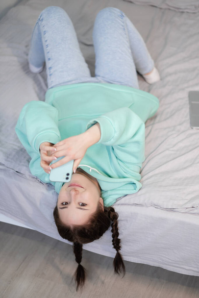 vzdálená práce nebo vzdálené studium z domova. mladá brunetka žena dívka blogger na posteli s tyrkysové moderní telefon. karanténní omezení. covid-19 pandemie - Fotografie, Obrázek