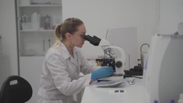 Női egészségügyi szakember dolgozik minták és elemzések orvosi laboratóriumban mikroszkóp mögött. Női labor technikus elemzi a mintát mikroszkóppal a kórházban. Gyógyászat, mikrobiológia - Felvétel, videó
