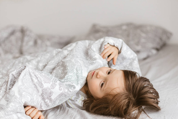 An adorable little boy plays around on the bed and wraps himself in a blanket. Time with children at home. Childrens games during the holidays - Valokuva, kuva