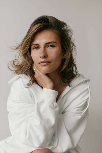Portrait of a beautiful young woman with brown hair and clear bronze skin without makeup on a white background. Natural and healthy look. Neutral interior. Soft light. Oversized white hoodie. Health and self care - Fotografie, Obrázek