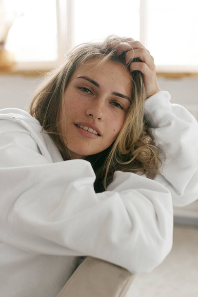 Portrait of a beautiful young woman with brown hair and clear bronze skin without makeup on a white background. Natural and healthy look. Neutral interior. Soft light. Oversized white hoodie. Health and self care - Fotoğraf, Görsel