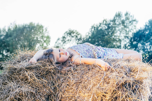 Portrait of little blond girl in dress alone lying and relaxing on haystack in field. Side view. Trees and sky on back - Fotó, kép