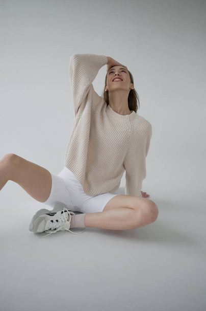 Cool sporty girl in beige sweater and white bicycle pants on neutral grey background. Soft light, monochrome picture. Good vibes, positive emotions. Tall and slim european model in good shape. - 写真・画像