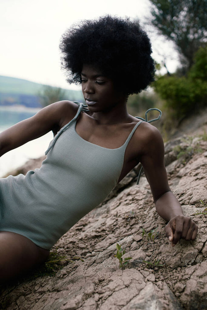 African beautiful model with perfect body in cerulean bodysuit. Voluminous hair, chocolate skin. Healthy looking. Water on background, natural colors. Summer landscape. Calm and balance.  - Foto, imagen