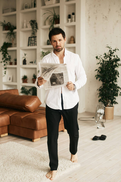 European man in his flat. Sitting in the living room. Bronze skin. Dark eyes and hair. Beard. Light colored interior. Casual clothes style. Weekend at home. Domestic atmosphere. Cosiness and comfort - Фото, изображение