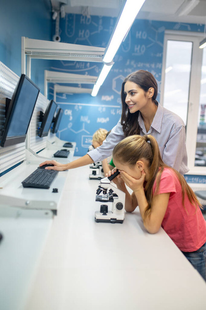 Frau blickt auf Bildschirm und Mädchen lehnt sich über Mikroskop - Foto, Bild