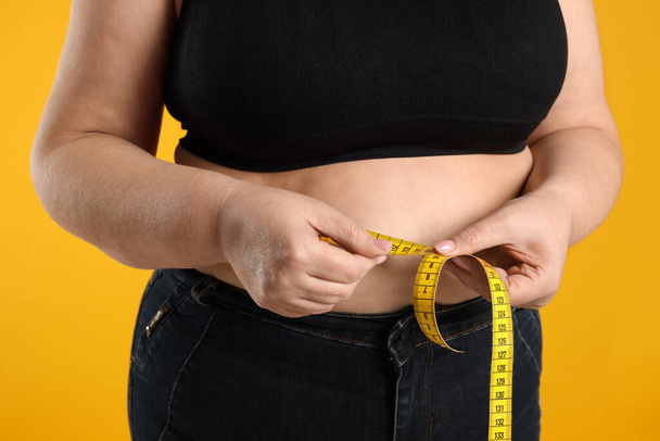 Overweight woman measuring waist with tape on orange background, closeup - Valokuva, kuva