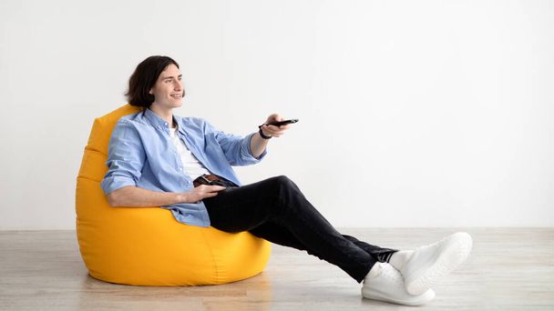 Watching TV. Young man resting in beanbag chair, switching channels with remote controller and looking at empty space - Fotografie, Obrázek