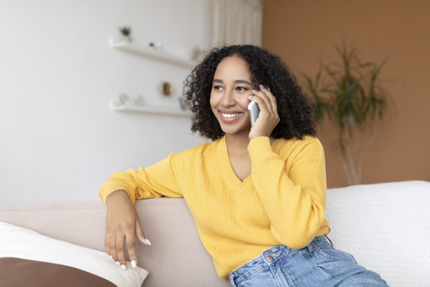 Vidám fiatal afro-amerikai nő beszél okostelefonon, ül a kanapén otthon - Fotó, kép