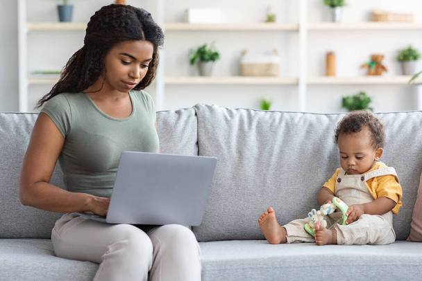 Молодая черная мама работает в Интернете за компьютером с маленьким ребенком, играя вокруг - Фото, изображение