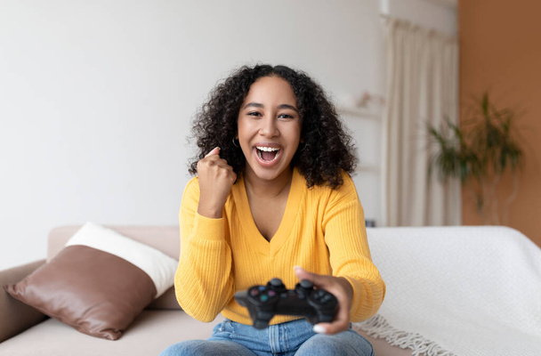 Emocionada joven afroamericana con joystick haciendo el gesto SI, ganando el videojuego, sentada en el sofá en casa - Foto, Imagen
