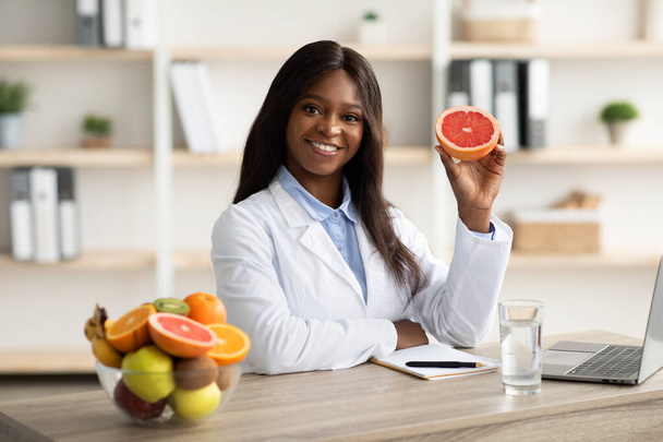 フレンドリーな黒の女性の栄養士は、グレープフルーツを保持し、カメラで笑顔、オンライン減量の相談を提供 - 写真・画像