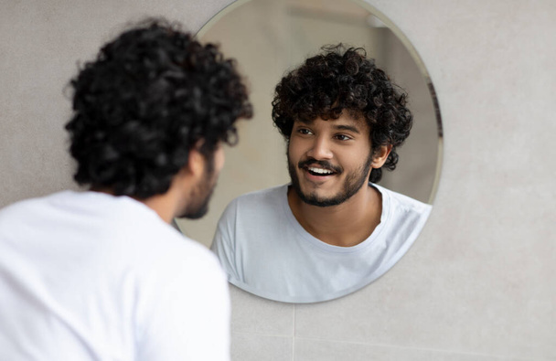 Portrait of happy indian man looking at his reflection in round mirror on the wall, young guy standing in bathroom - Foto, afbeelding