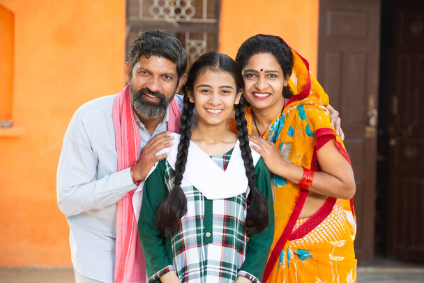 Portrait of happy Indian family standing outside their house in village Smiling Parents with their adorable young daughter in traditional outfit. rural india concept. looking at camera - Photo, Image