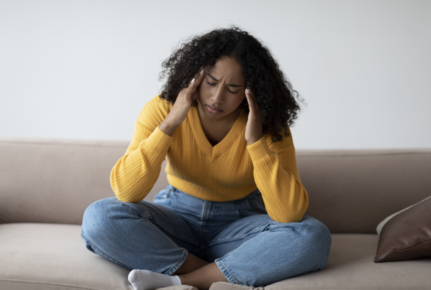 Молода чорна жінка з депресією торкається її храмів, відчуваючи стрес на дивані вдома
 - Фото, зображення