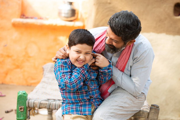 Ο νεαρός ευτυχισμένος ινδιάνος πατέρας που διασκεδάζει μαζί του είναι χαριτωμένος μικρός γιος. Γενειοφόρος που παίζει με το παιδί, Παιδί που γελάει, αγροτική Ινδία. - Φωτογραφία, εικόνα