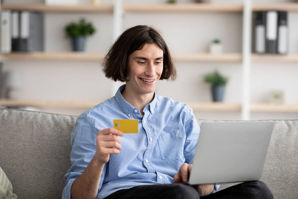 Шопинг и электронная торговля, молодой парень с кредитной картой, совершающий покупки онлайн через ноутбук, сидящий дома на диване - Фото, изображение