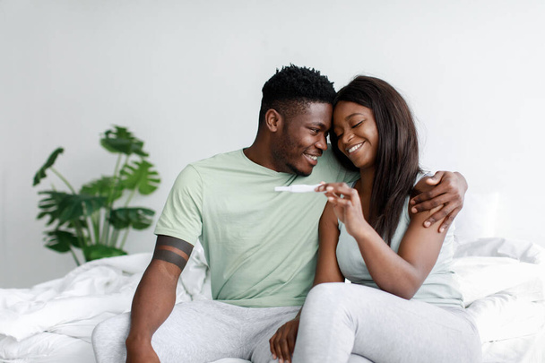 Glad happy young african american lady and guy look at pregnancy test on white bed in bedroom interior - Photo, Image