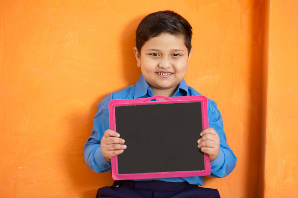Okul üniformalı mutlu Hintli bir çocuğun portresi turuncu arka planda boş bir sayfa tutuyor, sevimli ilkokul çocuğu kara tahta gösteriyor. Çocuk eğitimi konsepti. kırsal Hindistan. - Fotoğraf, Görsel