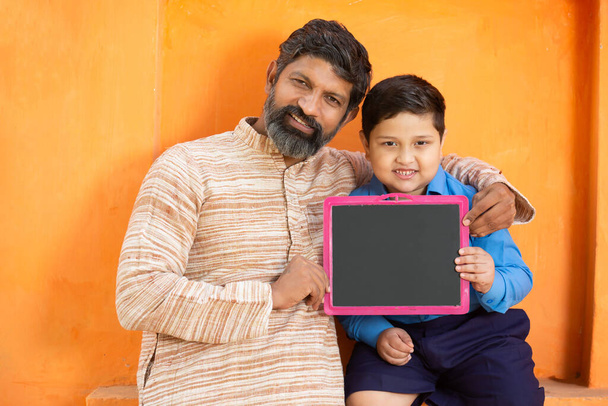 Pai indiano feliz e seu filho bonito em uniforme escolar segurando ardósia em branco contra fundo laranja, garoto elementar adorável com barba homem mostrando placa preta. educação infantil, india rural. - Foto, Imagem