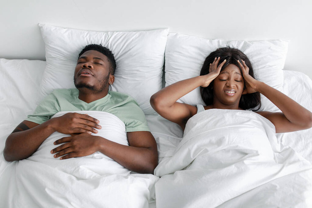 Λυπημένη απελπισμένη νεαρή Αφροαμερικανή σύζυγος που υποφέρει από πονοκέφαλο, θόρυβο και ροχαλητό κοιμισμένο σύζυγο στο κρεβάτι - Φωτογραφία, εικόνα