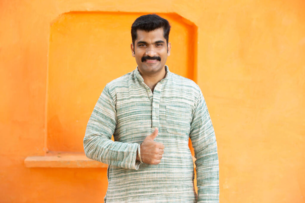 Портрет счастливого индийского усатый мужчина в традиционной курта сделать большие пальцы вверх поза стоя на оранжевом фоне, крупным планом глядя в камеру. - Фото, изображение