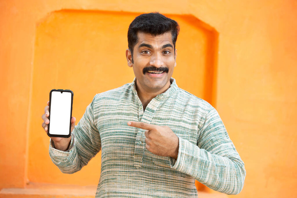 Cheerful Indian young mustache man showing blank phone screen , excited Male point finger display empty smart phone for advertisement and branding over orange background, mockup. - Photo, Image
