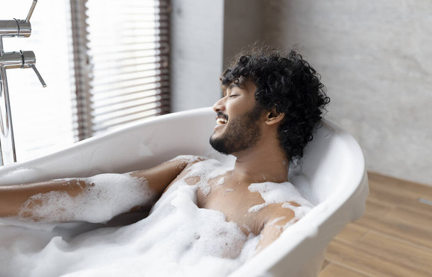 Расслабляющая ванна. Индийский мужчина лежит в ванне, наслаждаясь пеной на дому спа, отдых в горячей воде дома, свободное место - Фото, изображение
